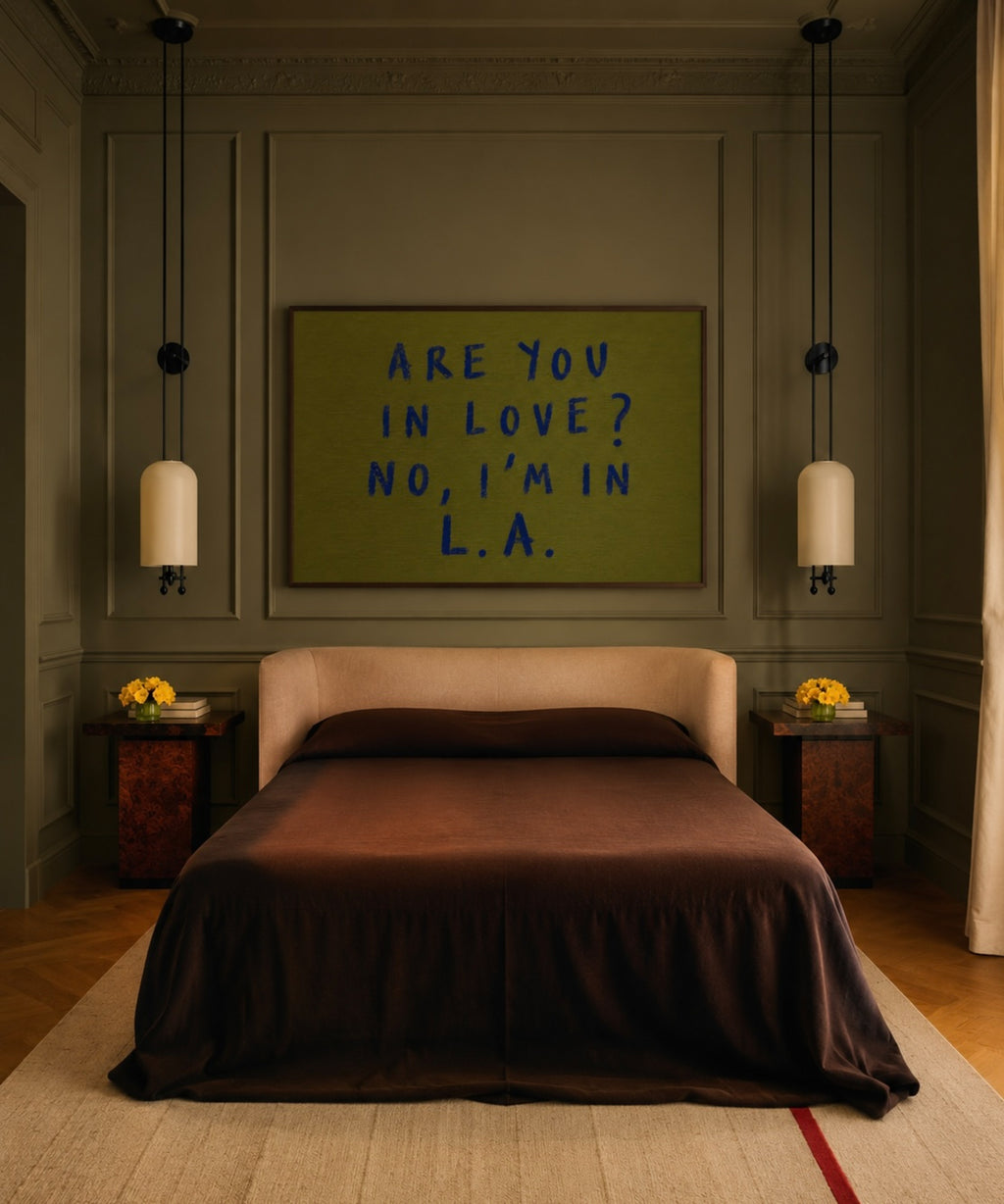 A styled bedroom interior featuring a bed with dark brown bedding, flanked by matching bedside tables and pendant lights, with a framed art print on the wall above the headboard that reads 'ARE YOU IN LOVE? NO, I'M IN L.A.