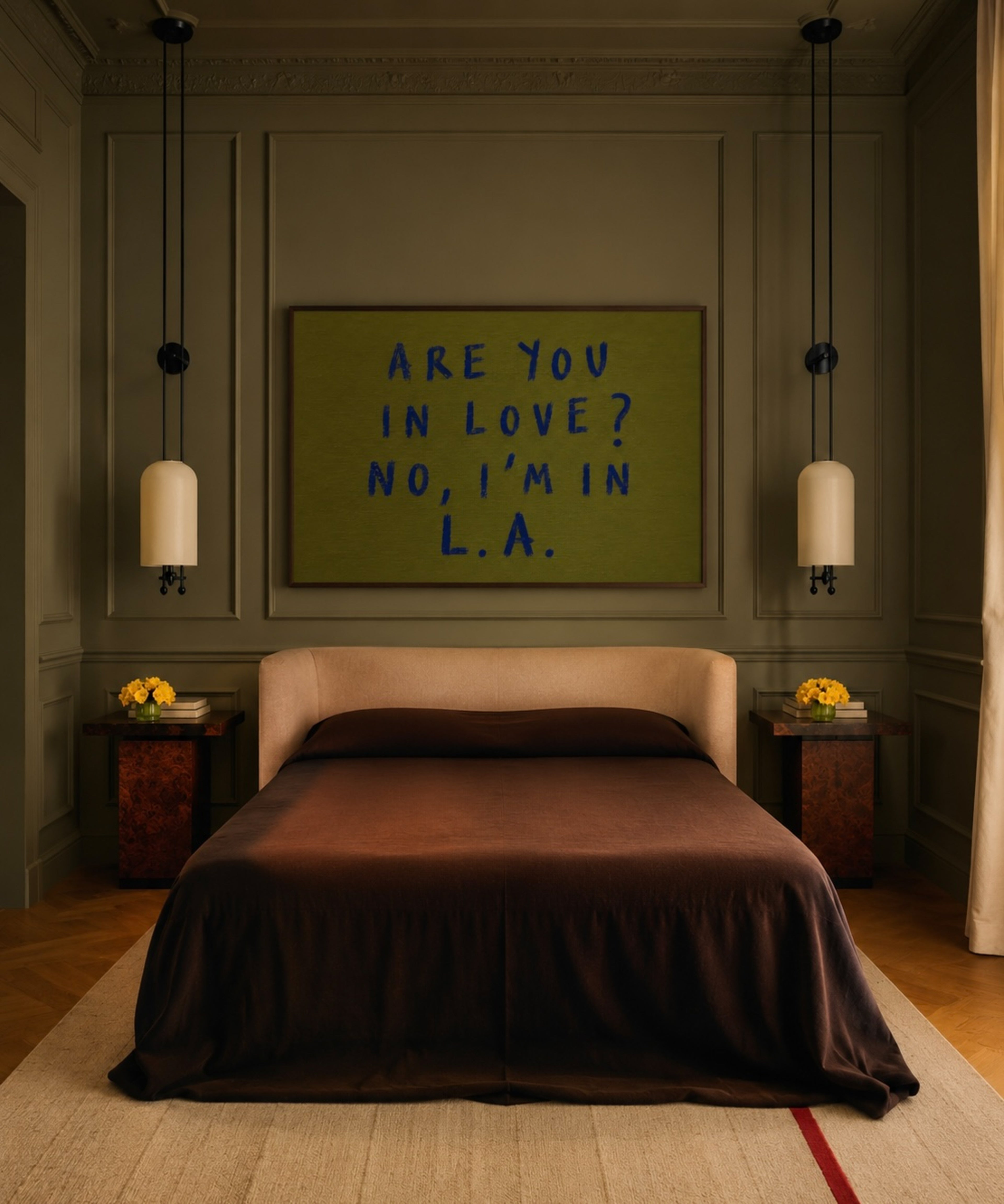 A styled bedroom interior featuring a bed with dark brown bedding, flanked by matching bedside tables and pendant lights, with a framed art print on the wall above the headboard that reads 'ARE YOU IN LOVE? NO, I'M IN L.A.