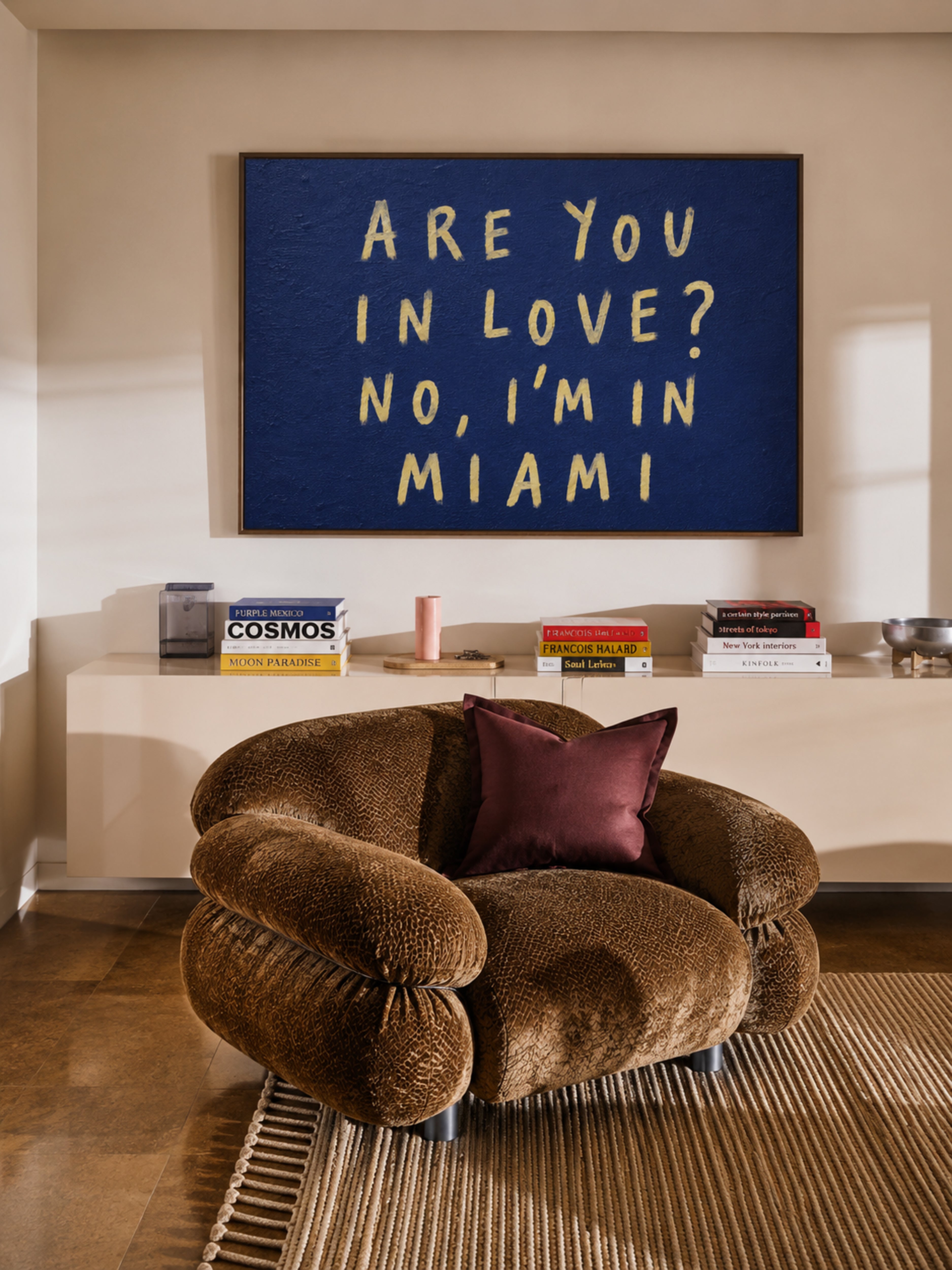 A cozy living room scene featuring a brown textured boucle armchair with a dark purple pillow, set in front of a cream console table. Above the table hangs a navy blue framed canvas print with cream-colored text that reads 'ARE YOU IN LOVE? NO, I'M IN MIAMI'.