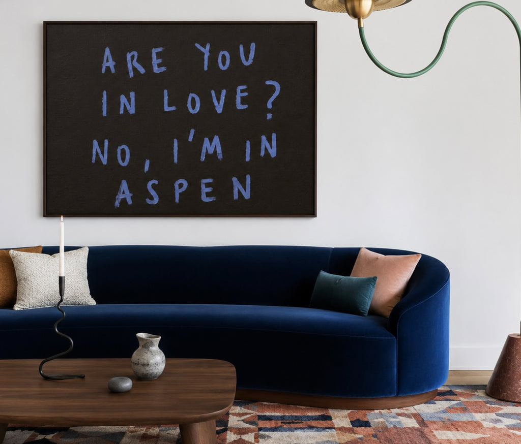 A framed wall art print with the text 'ARE YOU IN LOVE? NO, I'M IN ASPEN' in light blue lettering on a dark charcoal background, displayed above a navy blue velvet sofa in a modern living room setting.
