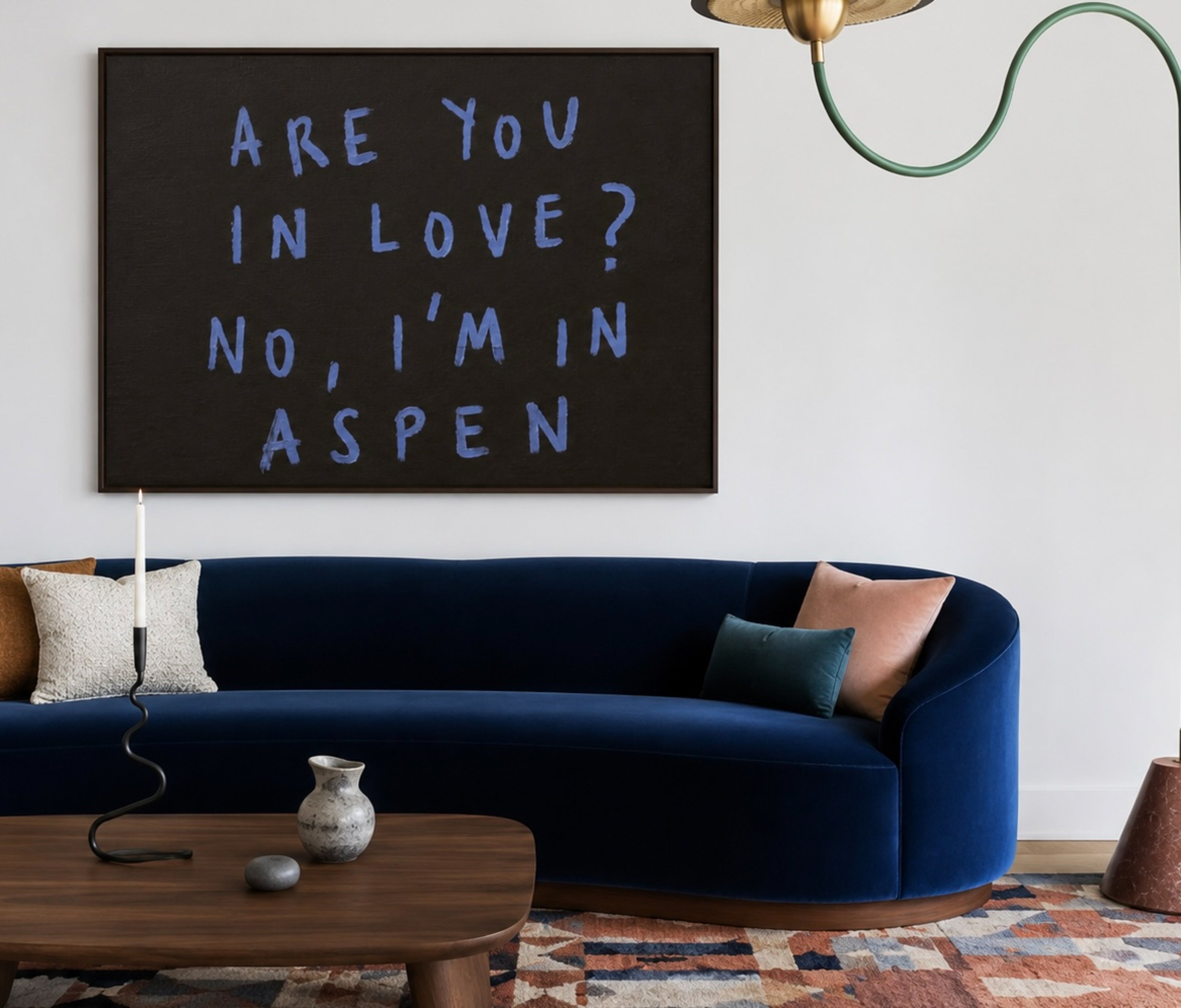 A framed wall art print with the text 'ARE YOU IN LOVE? NO, I'M IN ASPEN' in light blue lettering on a dark charcoal background, displayed above a navy blue velvet sofa in a modern living room setting.