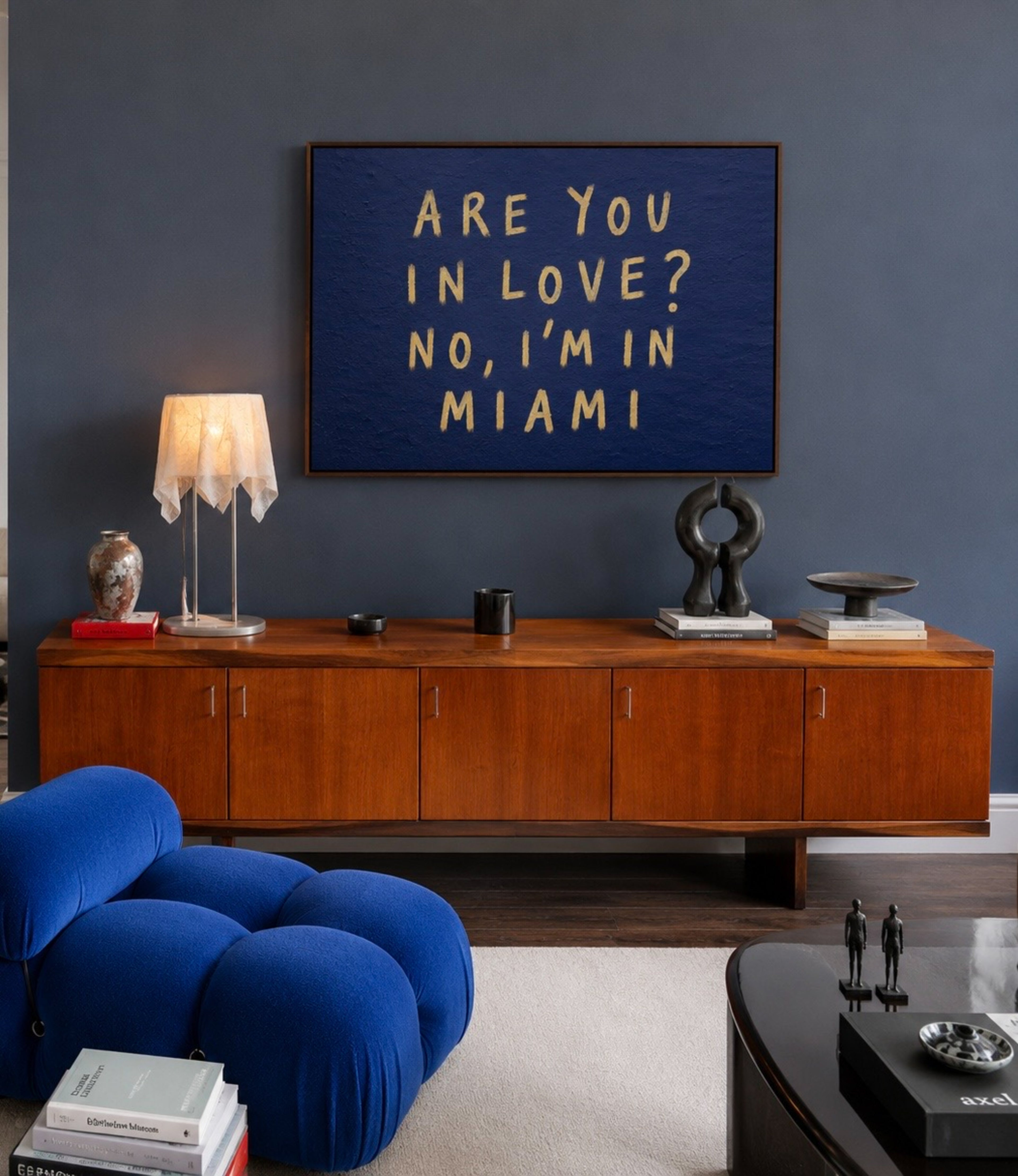 A framed square wall art print with the phrase 'ARE YOU IN LOVE? NO, I'M IN MIAMI' in gold lettering on a dark blue textured background, displayed above a mid-century modern wooden credenza in a styled living room.