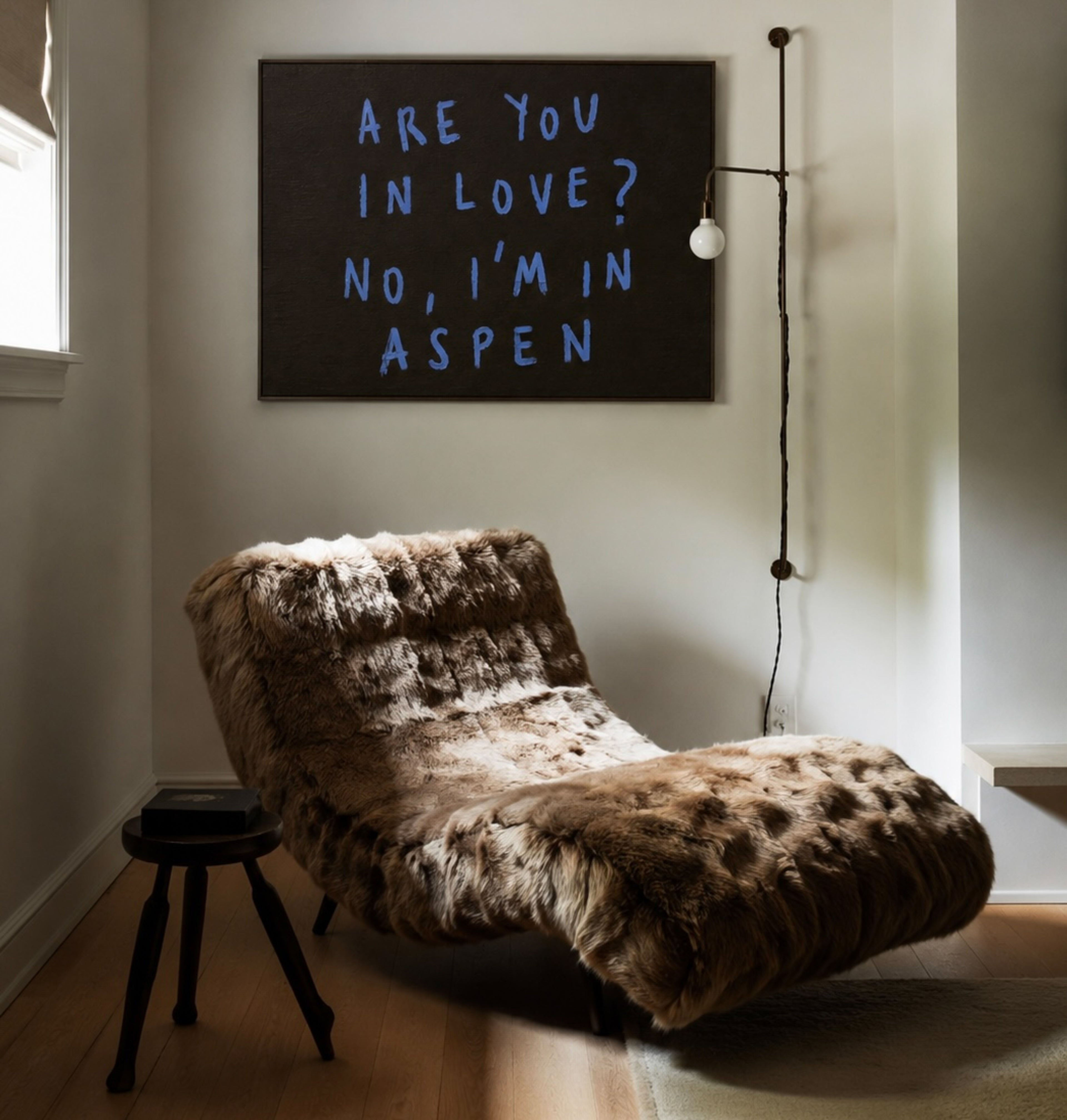 A rustic faux-fur chaise lounge chair in brown and beige tones, styled in a minimalist room with a black chalkboard wall sign that reads 'ARE YOU IN LOVE? NO, I'M IN ASPEN' in blue chalk.