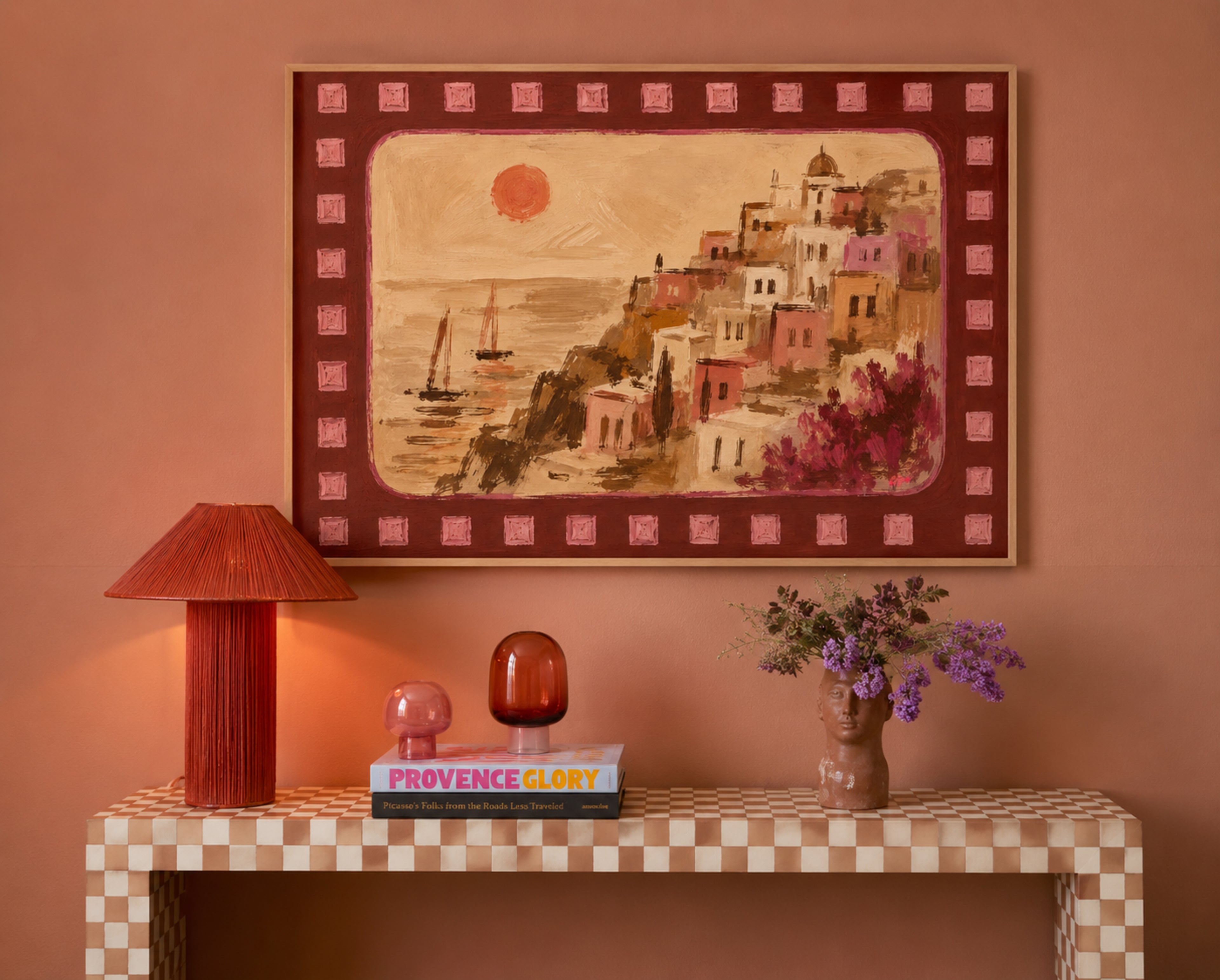 A framed art print depicting a coastal hillside village with sailboats under a red sun, displayed on a terracotta wall above a checkered console table with a red lamp and vase of purple flowers.