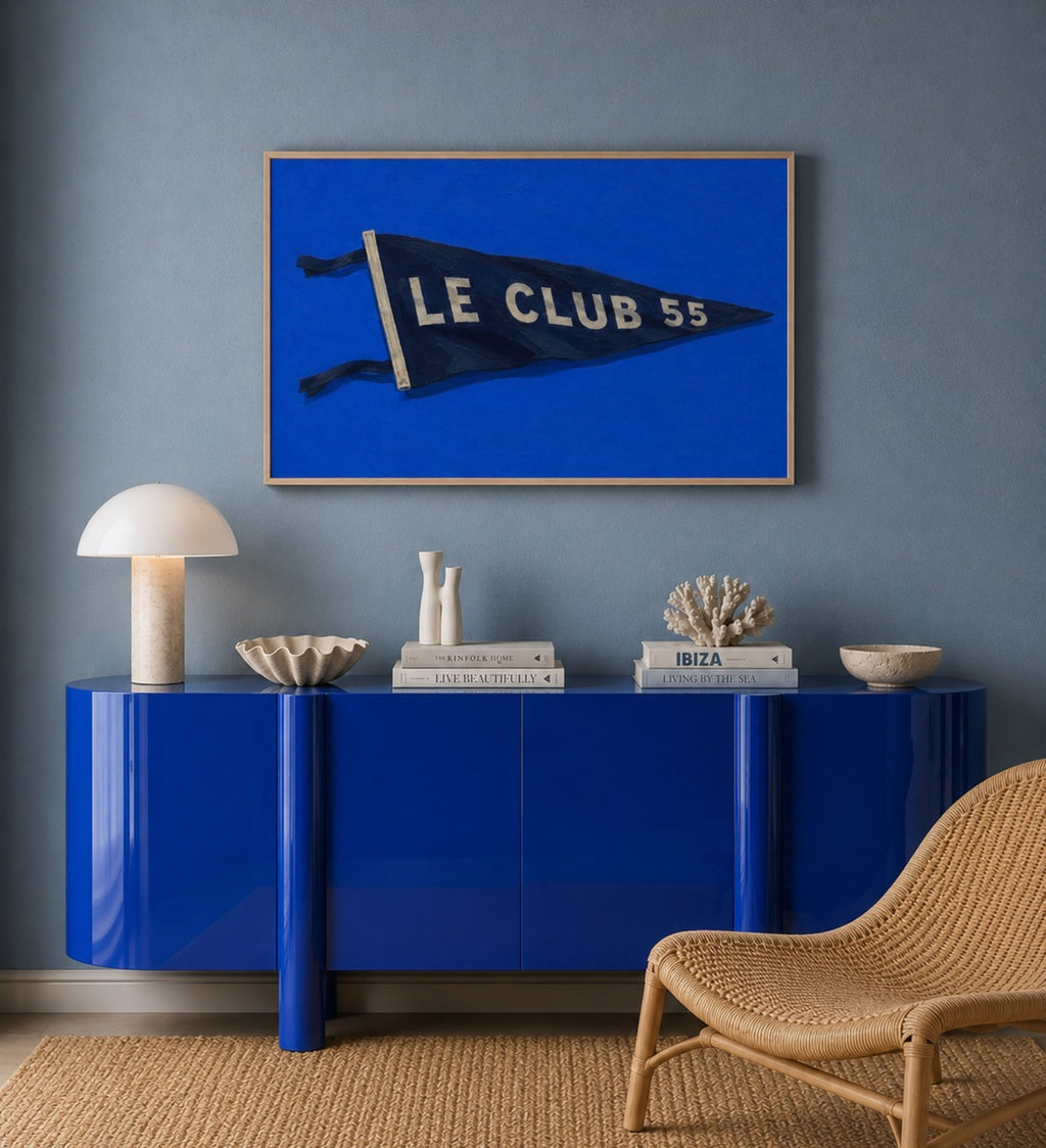 A framed art print featuring a vintage blue football pennant with 'LE CLUB 55' in white, displayed on a textured blue wall above a glossy blue sideboard with decorative objects and a woven chair.