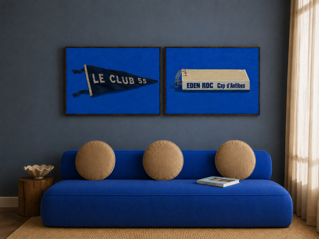 A styled interior scene with a blue sofa, three round beige cushions, a small side table, and two framed blue wall art prints hanging above the couch. The prints feature vintage football club graphics, including a black pennant reading 'LE CLUB 55' and a white box with 'EDEN RDC Cup d'Antibes'.