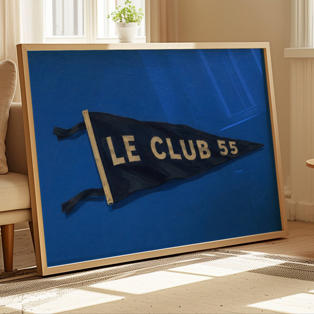 A framed art print leaning against a beige sofa in a sunlit living room. The print shows a dark blue pennant flag with the words 'LE CLUB 55' in cream lettering on a vivid blue background.