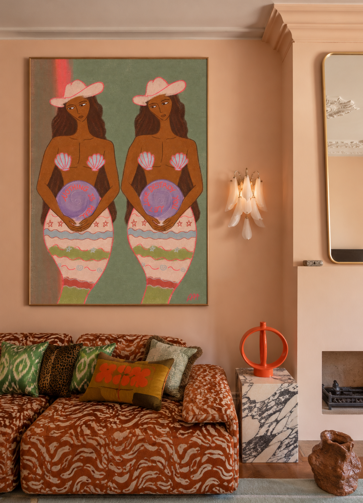 A large framed art print depicting two stylized mermaids in pink hats hangs on a peach-colored wall in a decorated living room with a patterned sofa and pillows.
