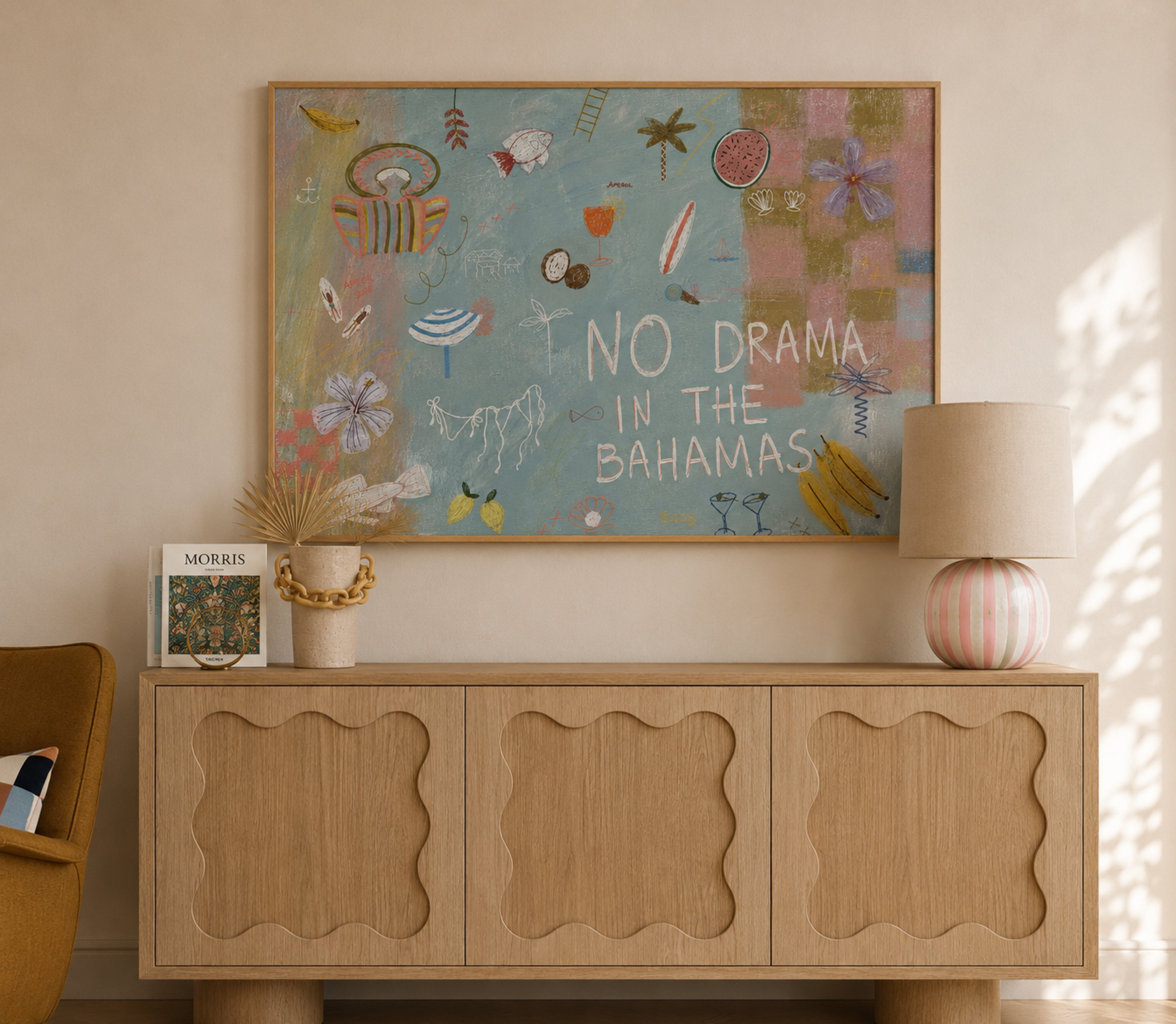 A framed art print with the text 'NO DRAMA IN THE BAHAMAS' hangs on a beige wall above a light wood console table. The print features a colorful tropical collage with various illustrations on a light blue background.