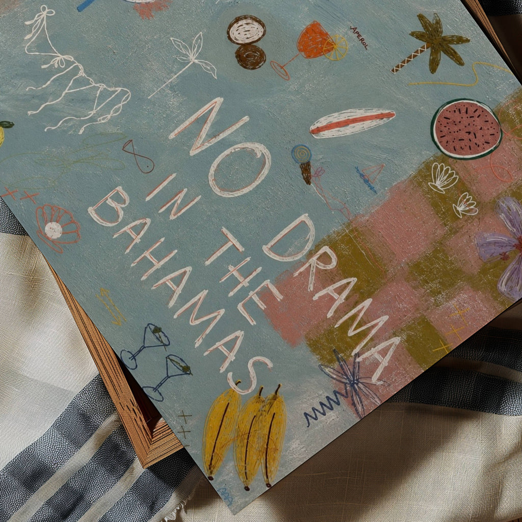 A square art print  featuring the text 'NO DRAMA IN THE BAHAMAS' in large white letters over a painted beach scene. The artwork includes tropical motifs like palm trees, a banana, watermelon, sunglasses, and flowers on a blue-green background, with pink sand and a striped blanket visible underneath.