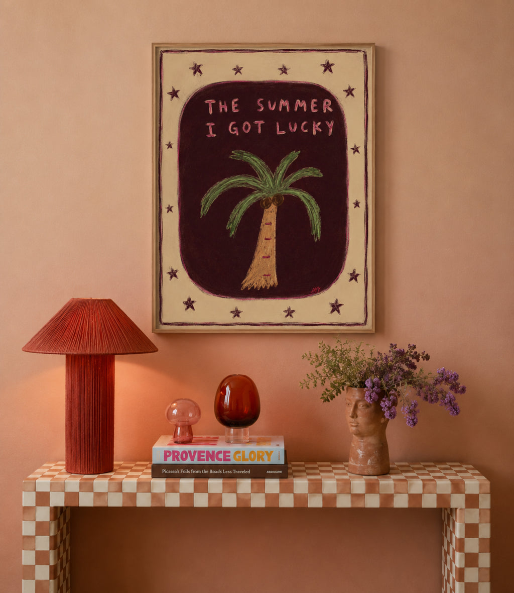 Framed wall art featuring a hand-drawn palm tree and the text 'THE SUMMER I GOT LUCKY' in pink, displayed on a peach wall above a checkered console table with a red lamp, books, and a vase of purple flowers.