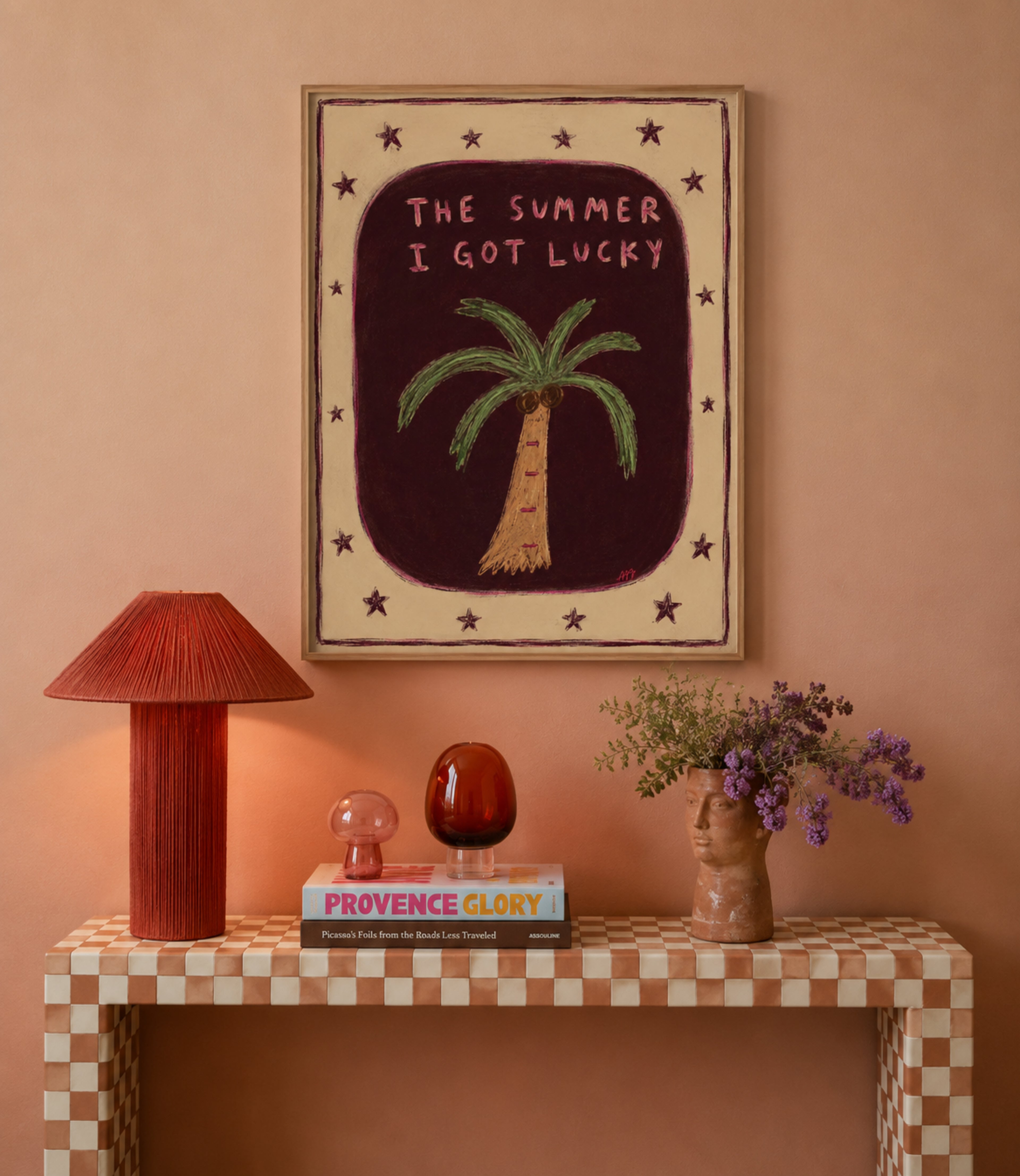 Framed wall art featuring a hand-drawn palm tree and the text 'THE SUMMER I GOT LUCKY' in pink, displayed on a peach wall above a checkered console table with a red lamp, books, and a vase of purple flowers.