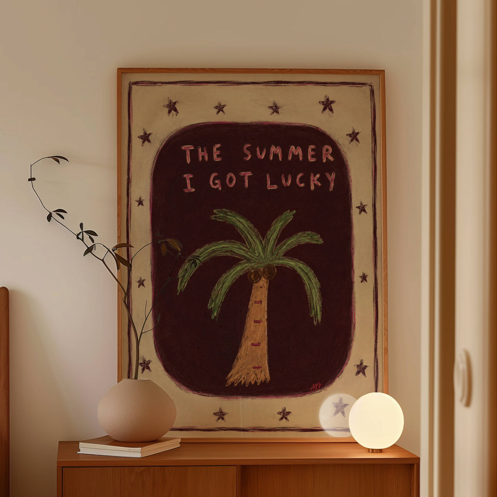 A framed art print with the text 'THE SUMMER I GOT LUCKY' and a palm tree illustration, displayed on a wooden console table in a warm, styled room setting.