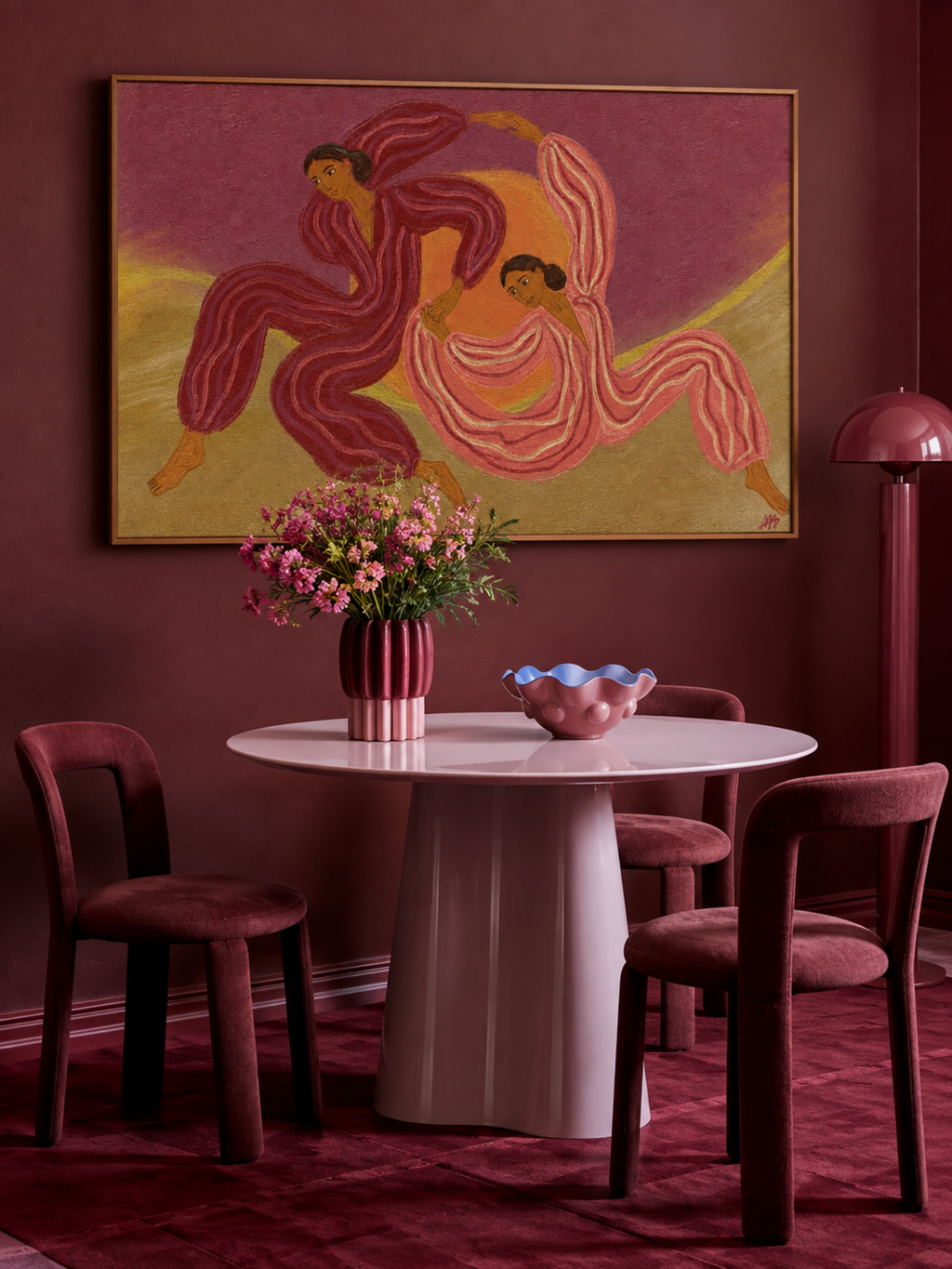 A styled dining room scene featuring a large framed art print of two dancing figures in pink and gold on a dark burgundy wall. Below the artwork is a round white table with a pink ribbed pedestal base, surrounded by four burgundy upholstered chairs.