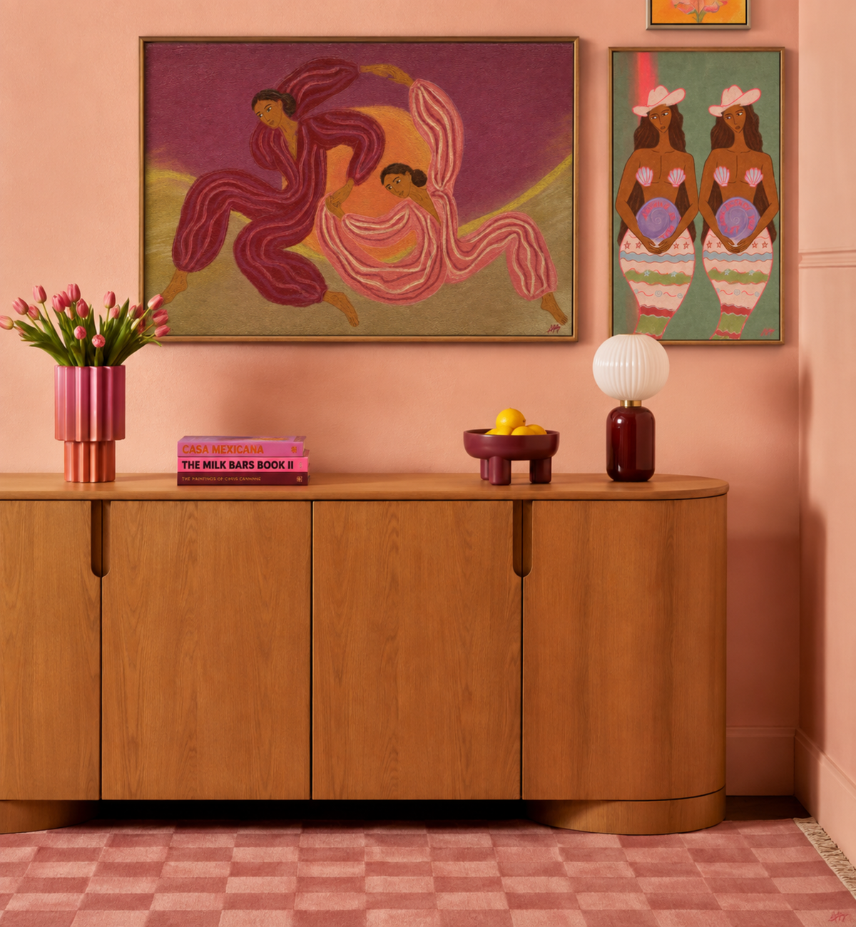 A styled interior scene with a curved wooden sideboard against a peach-pink wall, decorated with two framed art prints above. The left print shows two dancing figures in pink and purple tones, and the right print shows two figures in colorful striped garments.
