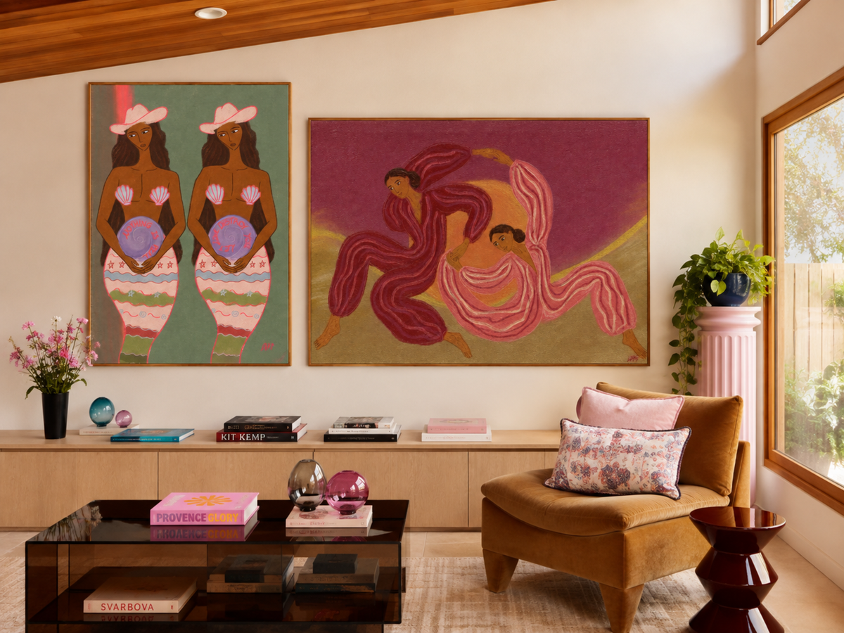 A styled interior room with two large framed art prints hanging on a beige wall above a low cabinet. The left print shows two women in pink hats and striped skirts on a green background, and the right print shows two women in flowing pink and magenta garments on a mauve and gold background.