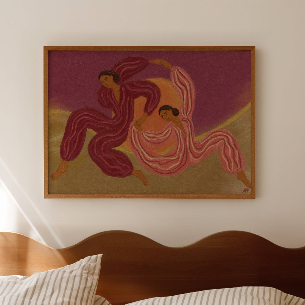 A framed art print depicting two stylized dancers in flowing pink and burgundy garments against a purple and beige background, hung on a light beige wall above a wooden headboard.