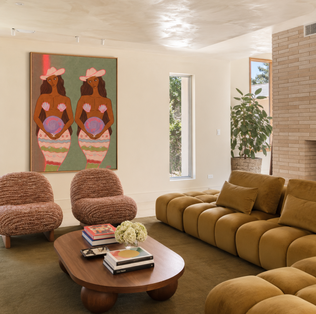A modern living room featuring a large framed art print of two women in pink hats on the wall, a mustard yellow modular sofa, two textured brown armchairs, and a round wooden coffee table.