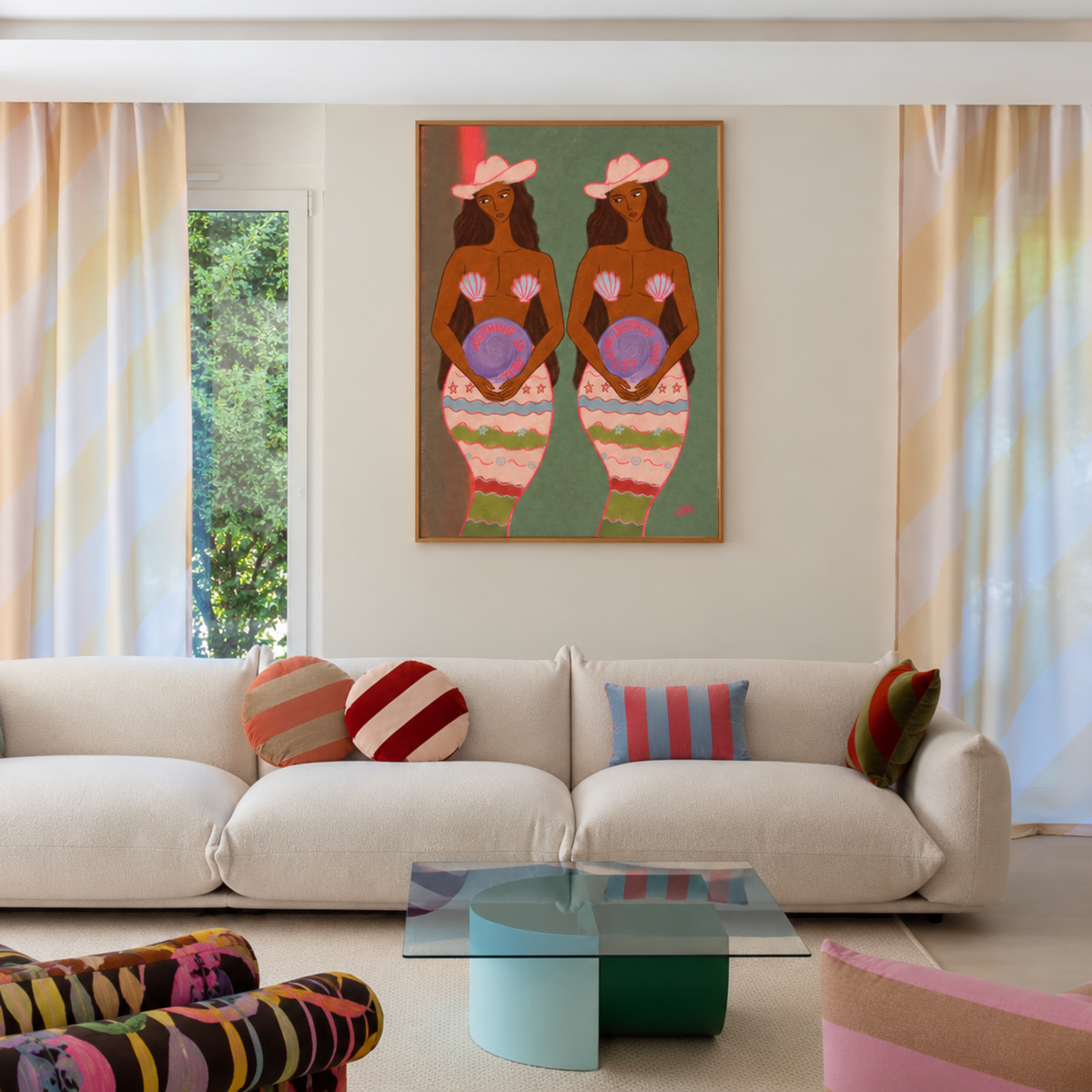 A colorful framed wall art print depicting two stylized figures with pink hats and patterned dresses, hung above a cream sofa in a bright, modern living room with striped curtains and colorful pillows.