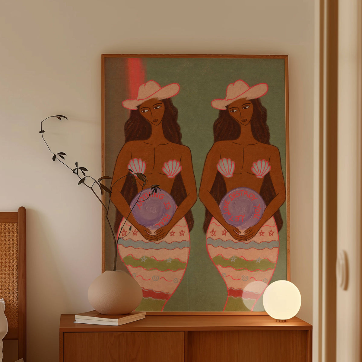 A framed art print featuring two stylized mermaid women with pink hats and patterned tails, displayed on a wooden console table in a warm, modern room with a vase and a glowing lamp.