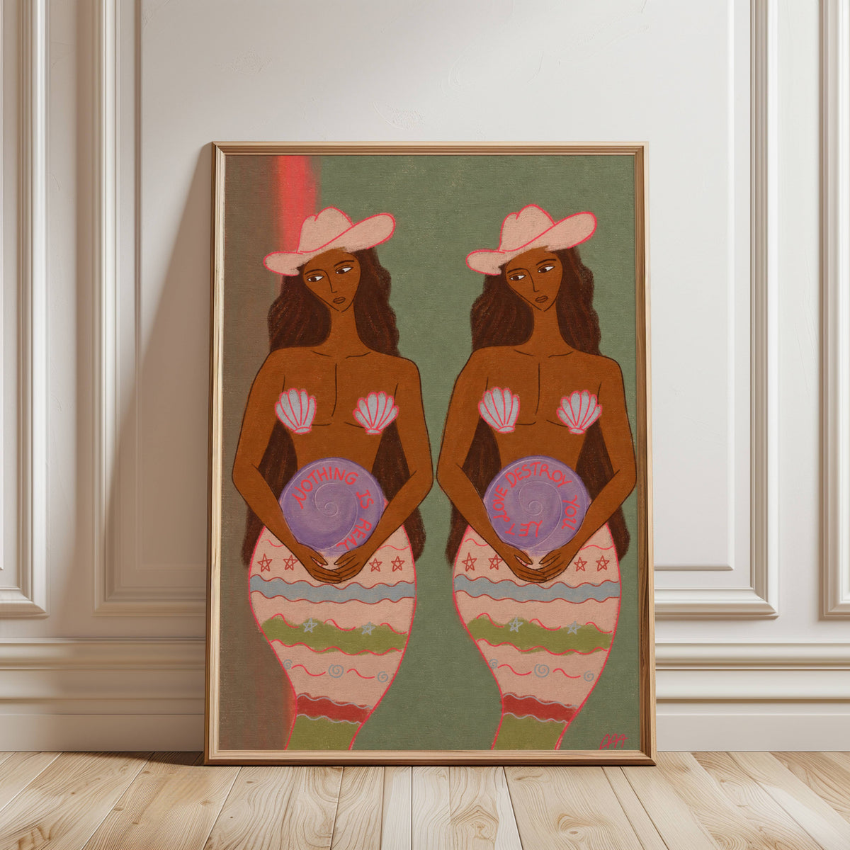 A framed art print leaning against a white paneled wall on a light wood floor. The artwork depicts two stylized Black women with pink hats and patterned mermaid-like lower bodies holding purple plates, set against a muted green background.