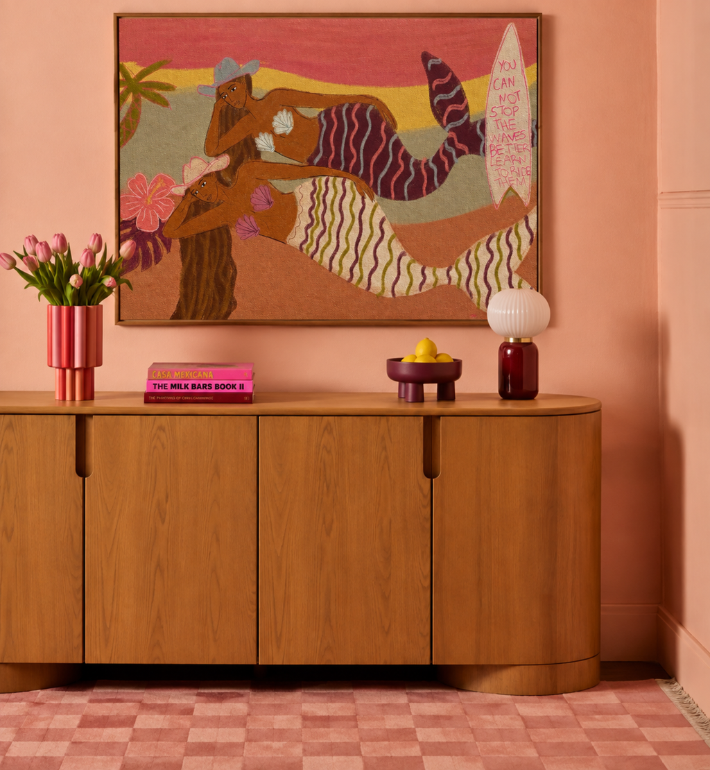 A framed colorful wall art piece depicting a stylized reclining figure with a striped tail and surfboard, displayed above a curved wooden sideboard in a pink-toned room with flowers, a lamp, and decorative objects.