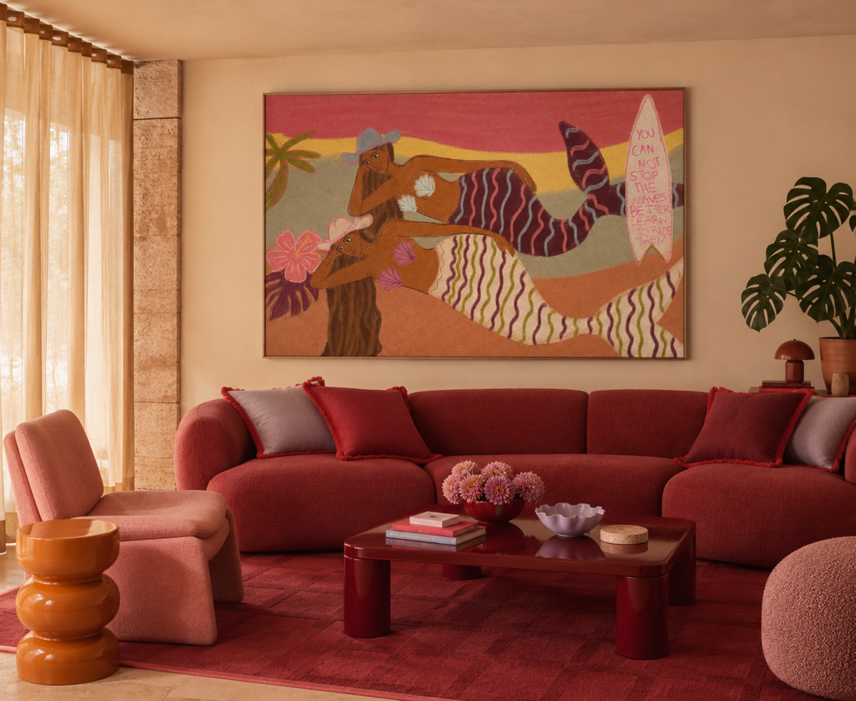 A large square wall art piece featuring a stylized beach scene with a woman, palm trees, and a surfboard, displayed above a red sectional sofa in a warm, modern living room.