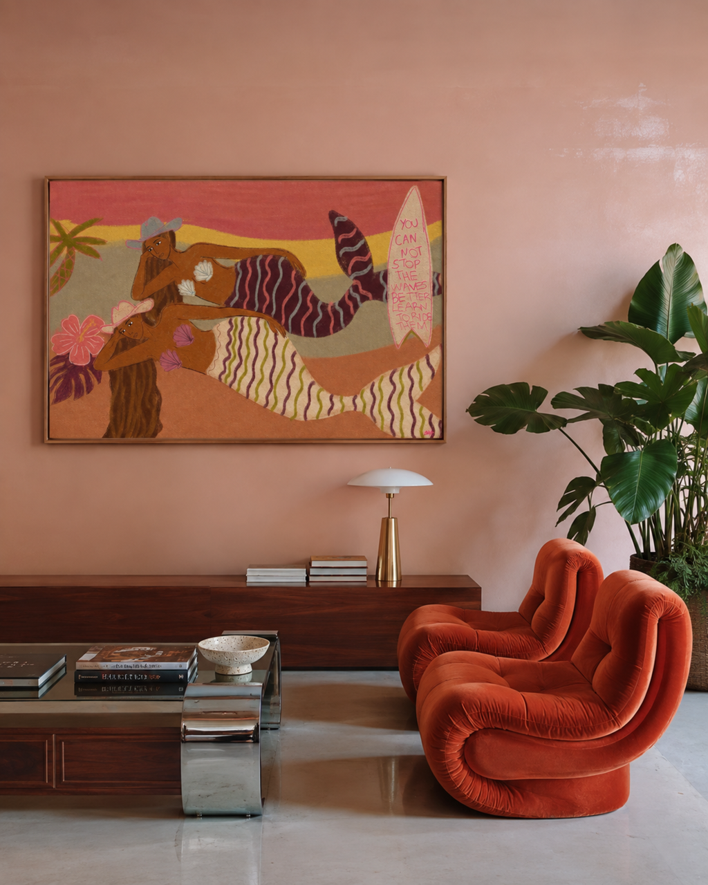 A bohemian-style framed wall art piece depicting a stylized figure and surfboard, displayed in a living room setting on a dusty pink wall above a wooden console table, next to a terracotta velvet armchair and a potted plant.