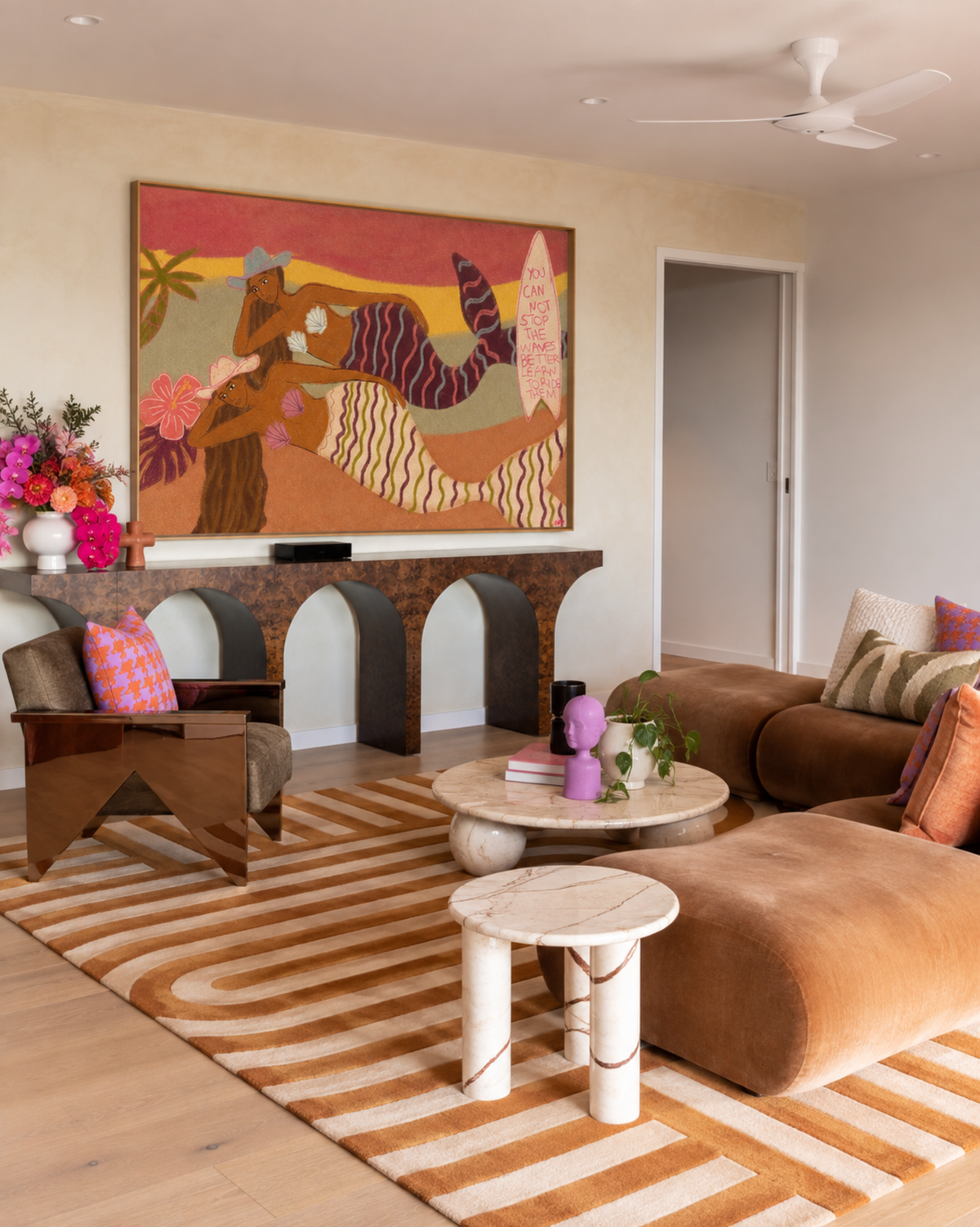 A large colorful abstract painting hangs on a beige wall above a decorative stone fireplace in a styled living room with a brown sectional sofa, striped rug, and small round tables.