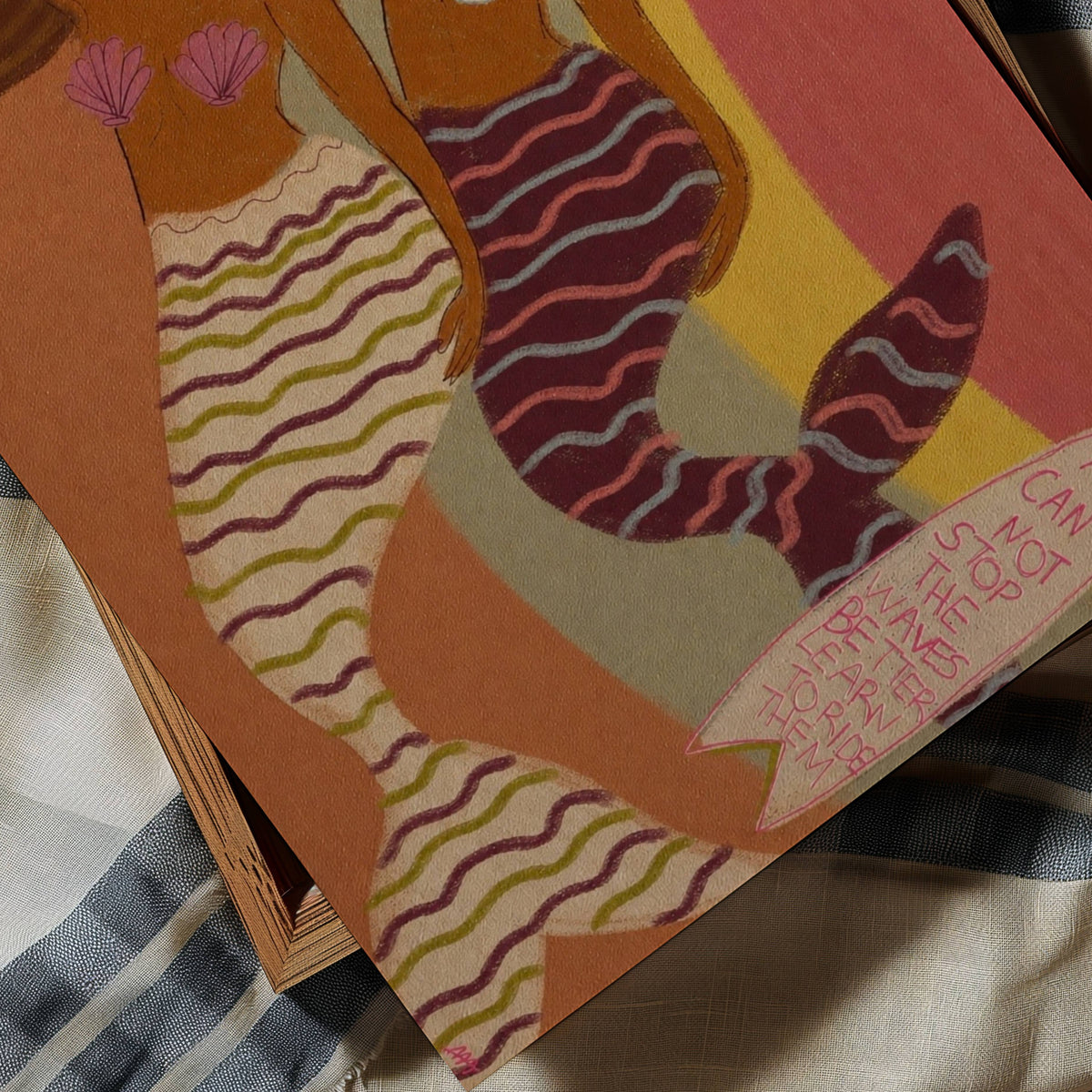 A close-up of a colorful art print featuring a stylized mermaid illustration, resting on a striped fabric surface.