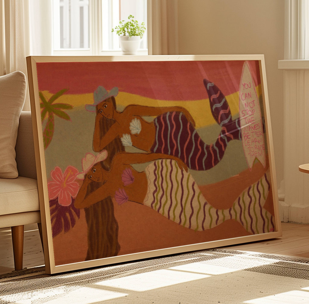 A large framed art print featuring two stylized mermaids in warm sunset tones, leaning against a sofa in a bright living room.