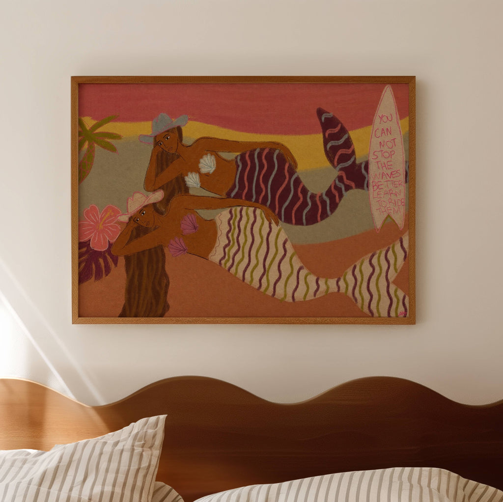 A framed art print featuring two stylized mermaids in a beach scene, hanging on a light beige wall above a wooden headboard with striped pillows.