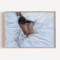 the art of staying is a hand painted wall art print of a sleeping womam in striped bedding, in light blue and butter yellow tones, inspired by slow mornings and nostalgic interiors, symbolizing quiet luxury and unhurried mornings. Horizontal above bed oil painting wall art print . sunday morning aesthetic intimate wall art