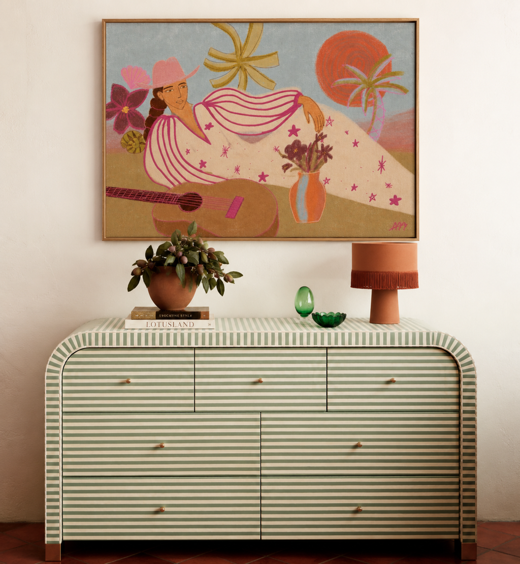 A framed art print featuring a retro-style illustration of a woman with a guitar and a sun, displayed on a wall above a striped cream and green dresser with a potted plant and a lamp.