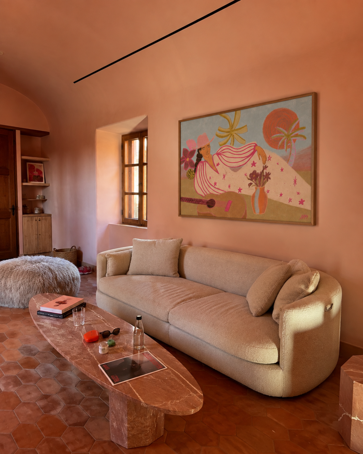 A large framed art print featuring a whimsical, retro-style illustration of a woman in a pink dress with palm trees and a sun, displayed on a terracotta-colored wall above a beige sofa in a rustic living room.