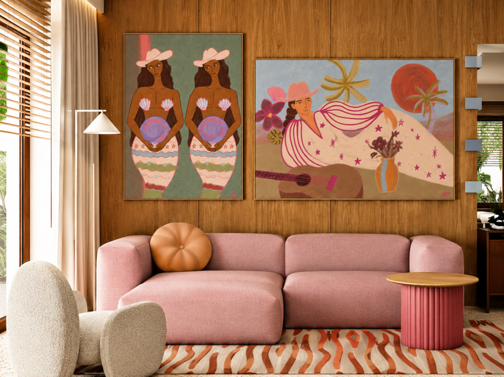 A styled interior room with a pink sectional sofa, wood-paneled wall, and two colorful framed art prints hanging above the couch. The left print shows two seated figures in pastel clothing, and the right print shows a seated figure with flowers, a guitar, and abstract shapes.
