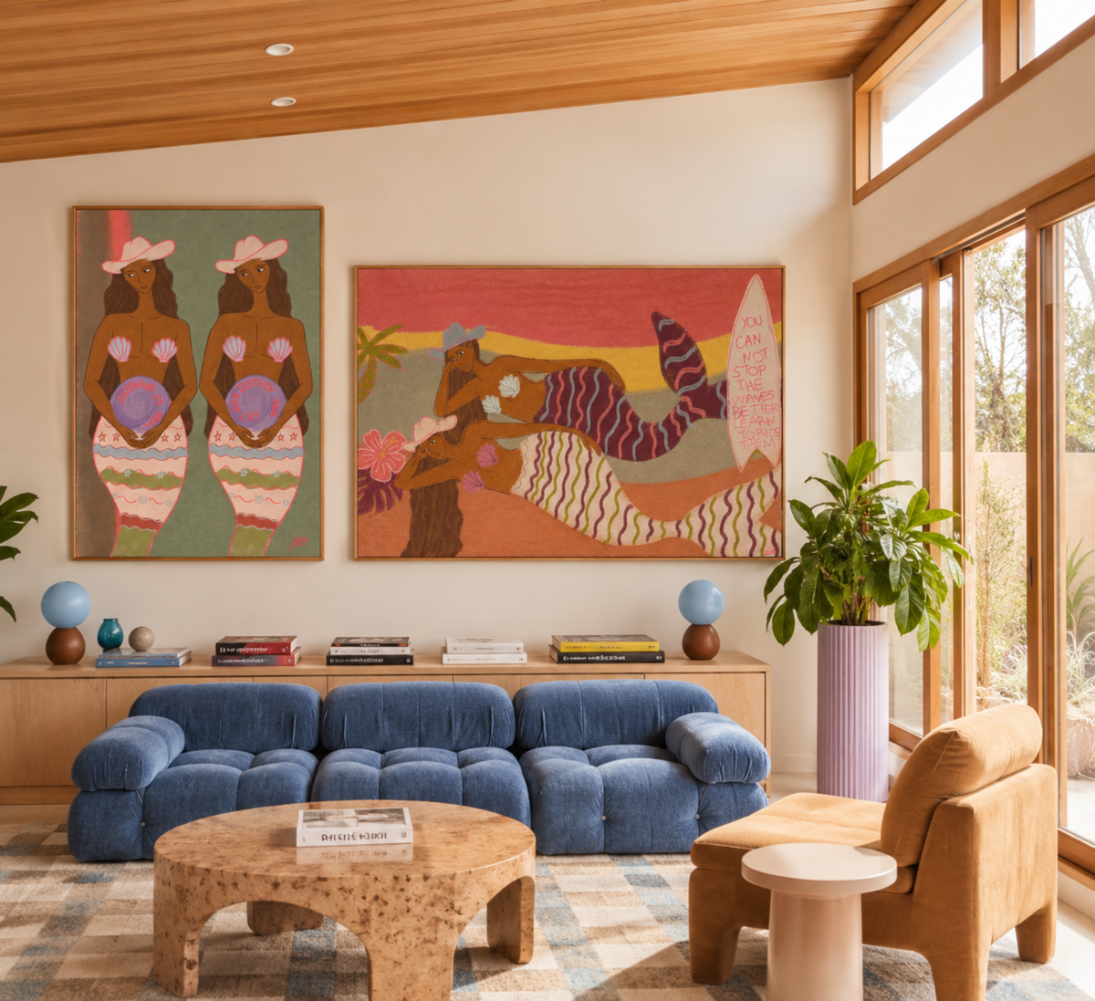 A styled living room interior with two colorful wall art pieces hanging above a blue sectional sofa. The artwork depicts stylized figures and a figure with a striped tail in warm earthy tones.
